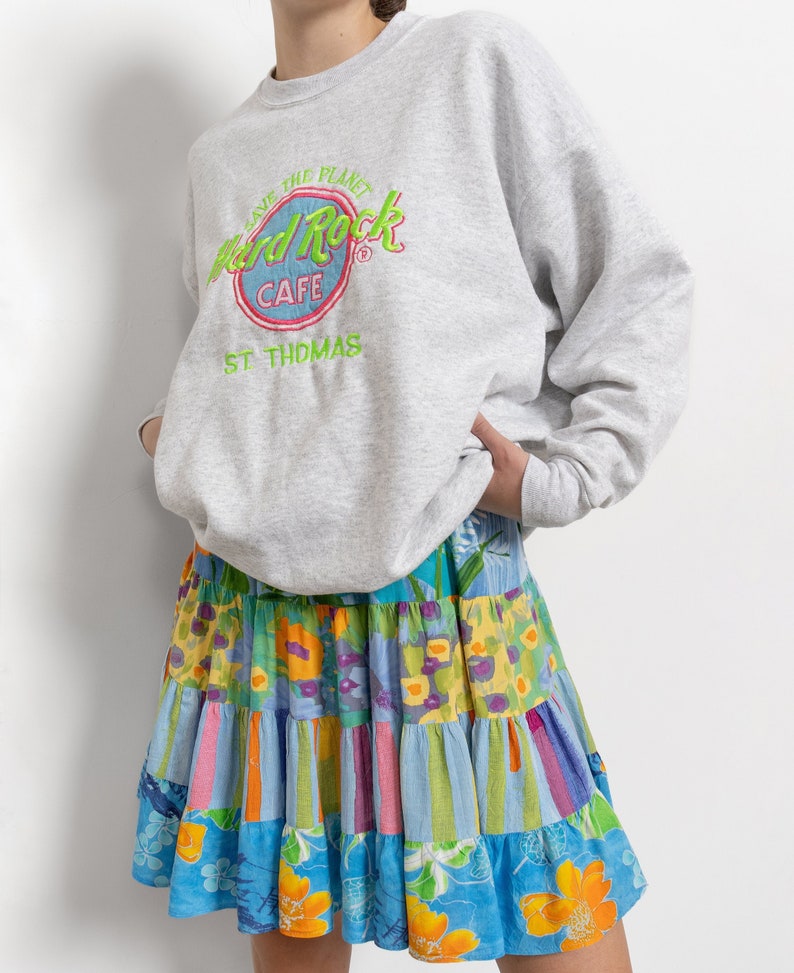 May include: A light gray crewneck sweatshirt featuring a Hard Rock Cafe St. Thomas logo with green and pink embroidery, paired with a tiered skirt showcasing a colorful patchwork pattern of floral and striped designs in blue, yellow, and pink.