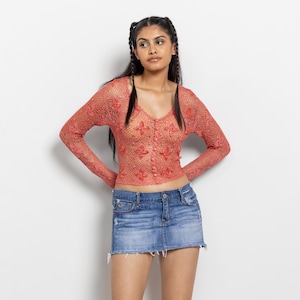 May include: A coral-colored long-sleeve crochet top featuring a V-neckline, a front button-down closure, and a repeating floral pattern, paired with a blue denim mini skirt that has a distressed finish and a frayed hemline at the bottom edge.