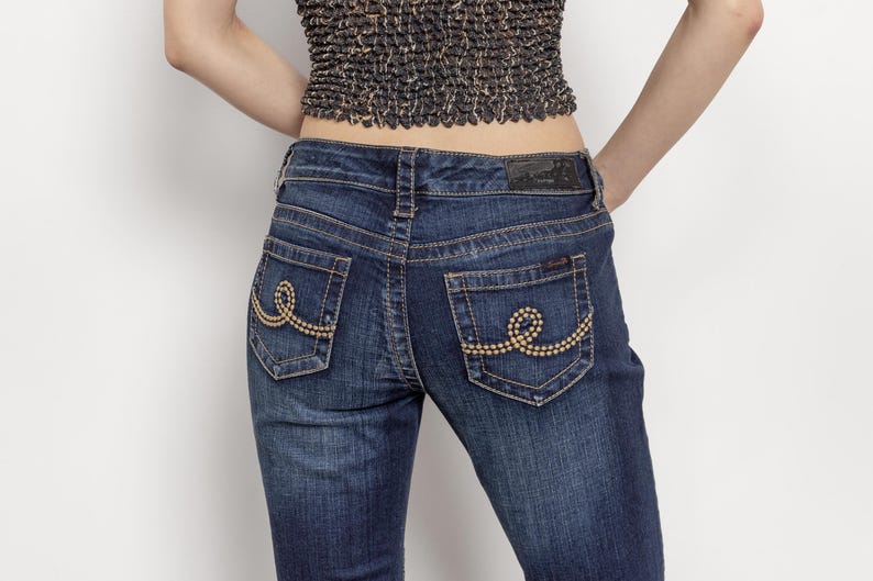 May include: Dark wash denim jeans with decorative stitching on the back pockets. The jeans have a classic five-pocket design and a dark leather patch on the waistband. The back pockets feature a gold-colored, beaded swirl design.