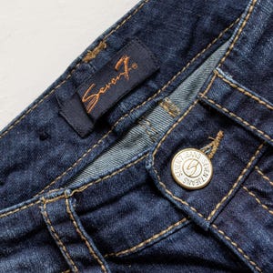 May include: Close-up of dark blue denim jeans. The jeans feature a button closure with the words "Seven Jeans" and a logo. The brand label is visible, and the stitching is in a contrasting tan color.
