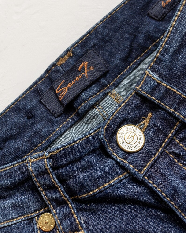 May include: Close-up of dark blue denim jeans. The jeans feature a button closure with the words "Seven Jeans" and a logo. The brand label is visible, and the stitching is in a contrasting tan color.