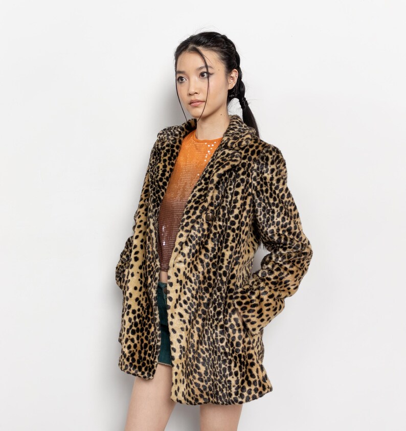 May include: A leopard print faux fur coat with a classic lapel collar. The coat features a brown and black spotted pattern. The model is wearing an orange sequined top and green shorts. The coat has pockets.