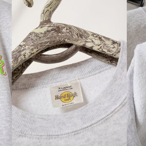 May include: A light gray crewneck sweatshirt featuring an embroidered Hard Rock Cafe St. Thomas logo in neon green, blue, and red. The front graphic reads Save The Planet Hard Rock Cafe St. Thomas. The interior tag indicates size X-Large.