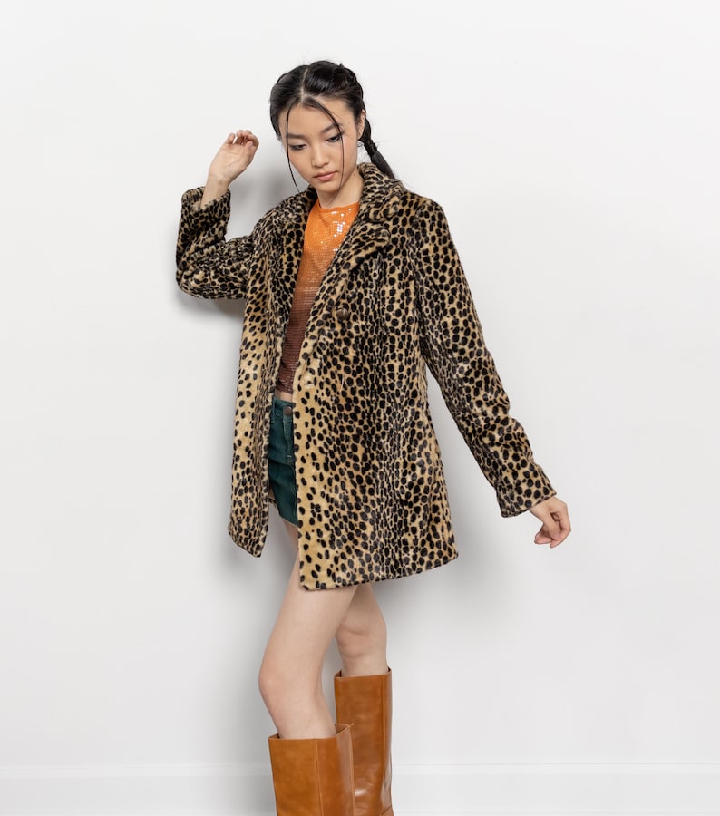 May include: A long, leopard-print coat with a classic collar and button closure. The coat features a brown and black spotted pattern. The model is wearing a brown top, green shorts, and brown boots. The background is a plain white wall.