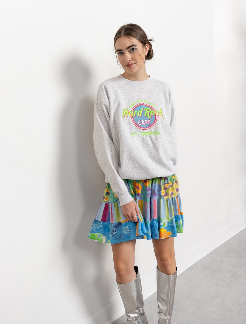 May include: A light gray crewneck sweatshirt featuring a Hard Rock Cafe St. Thomas logo in neon green and pink, paired with a multi-colored tiered mini skirt with floral and striped patterns, and metallic silver knee-high boots.
