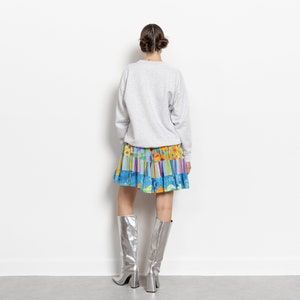 May include: A light gray long-sleeve crewneck sweatshirt paired with a short, tiered skirt featuring a vibrant multicolored floral and striped pattern, worn with knee-high metallic silver boots with a block heel and a square toe.