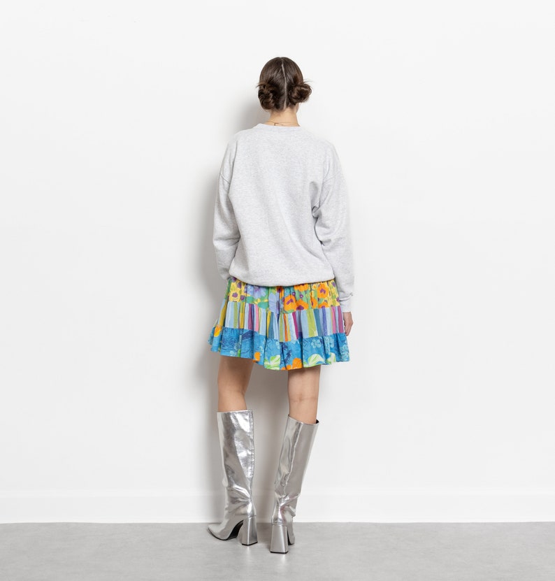 May include: A light gray long-sleeve crewneck sweatshirt paired with a short, tiered skirt featuring a vibrant multicolored floral and striped pattern, worn with knee-high metallic silver boots with a block heel and a square toe.