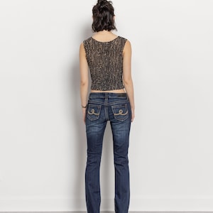 May include: A sleeveless, textured, brown and black top paired with dark wash blue jeans. The jeans have decorative stitching on the back pockets. The model is wearing maroon heels.