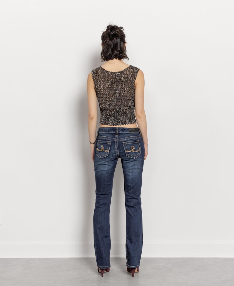 May include: A sleeveless, textured, brown and black top paired with dark wash blue jeans. The jeans have decorative stitching on the back pockets. The model is wearing maroon heels.