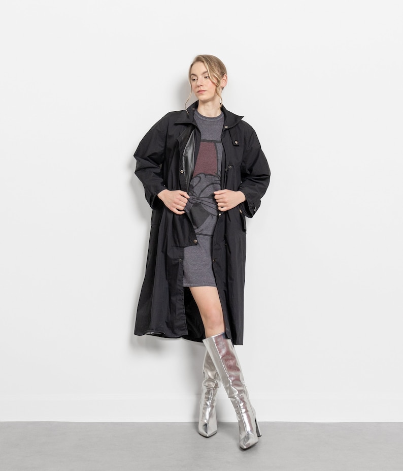Puede incluir: Un abrigo largo negro con cuello y botones. Se lleva sobre un vestido gris y se combina con botas plateadas hasta la rodilla. Las botas tienen punta y tacones altos. El conjunto se presenta sobre un fondo blanco.
