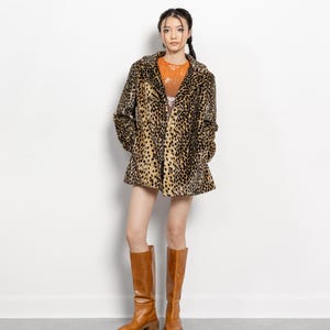 May include: A woman wearing a leopard print coat, orange top, and brown knee-high boots. The coat has a classic collar and a flared hem. The boots are a solid brown color with a chunky sole.