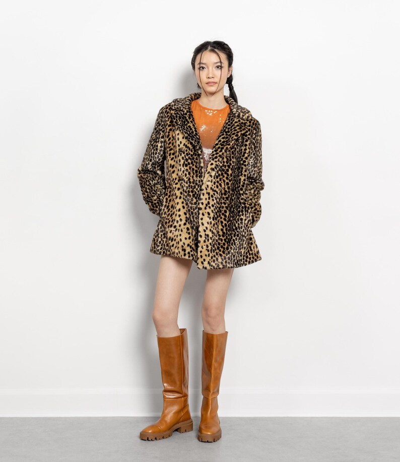 May include: A woman wearing a leopard print coat, orange top, and brown knee-high boots. The coat has a classic collar and a flared hem. The boots are a solid brown color with a chunky sole.