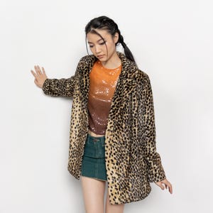 May include: A leopard print coat with a brown and black pattern. The coat is worn over an orange and brown sequined top and a green denim skirt. The coat has a classic lapel and a relaxed fit.