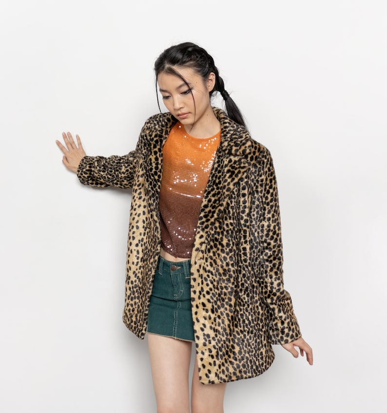 May include: A leopard print coat with a brown and black pattern. The coat is worn over an orange and brown sequined top and a green denim skirt. The coat has a classic lapel and a relaxed fit.