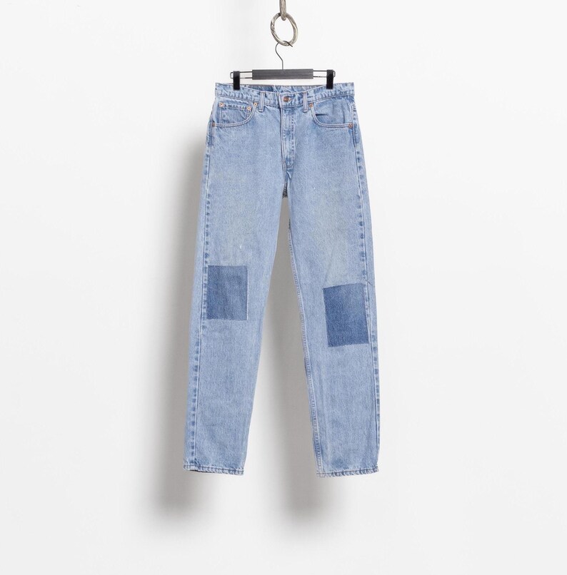 May include: Light blue denim jeans featuring a high-waisted cut, a classic five-pocket design, and a button fly closure. The legs of the jeans are accented with rectangular dark blue denim patches positioned over each knee area.