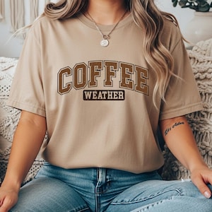 May include: A tan t-shirt with the words "COFFEE WEATHER" in brown block letters. The shirt is short-sleeved and has a crew neck. The model is wearing blue jeans.
