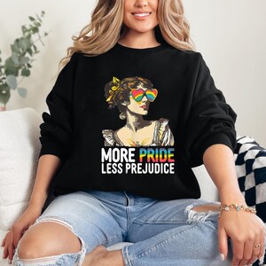 Op de afbeelding: Zwart sweatshirt met een afbeelding van een vrouw met regenboogkleurige hartvormige zonnebrillen. De tekst "MORE PRIDE LESS PREJUDICE" staat eronder in regenboogkleuren. Het sweatshirt heeft een casual stijl.