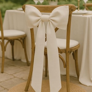 Op de afbeelding: Een houten stoel met een witte strik van stof die om de rugleuning is gebonden. De stoel staat aan een tafel met een wit tafelkleed en een boeket witte bloemen.