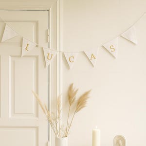 Puede incluir: Una guirnalda blanca con el nombre "LUCAS" en letras doradas cuelga sobre una mesa con un mantel blanco. Una vela blanca alta está sobre un posavasos de madera. Un jarrón blanco con flores secas está sobre un pedestal.