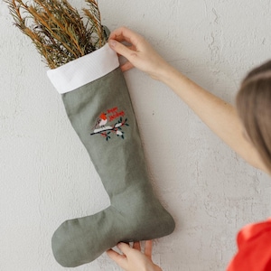 Könnte beinhalten: Ein salbeigrüner Weihnachtsstrumpf mit weißem Bündchen, gefüllt mit getrocknetem Laub. Der Strumpf hat ein gesticktes Design eines Vogels auf einem Ast mit den Worten "Happy Holidays".