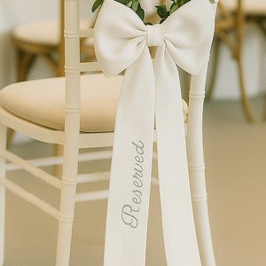 Op de afbeelding: Een witte stoel met een crèmekleurige zitting is versierd met een grote witte strik en groen. De lange linten van de strik zijn bedrukt met het woord "Reserved" in een scriptlettertype. De stoel maakt deel uit van een bruiloft of evenement.