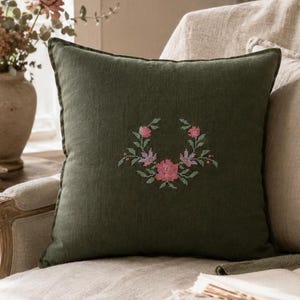 May include: A square, dark green decorative pillow with a floral cross-stitch design. The embroidered wreath features pink and purple flowers with green leaves. The pillow is on a beige upholstered chair.