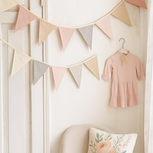 Op de afbeelding: Een zachtroze jurk met een gefranjerde zoom hangt aan een hanger. Twee slingers van pastelkleurige vlaggetjes versieren de muur. Een bloemenkussen ligt op een lichtgekleurde stoel.