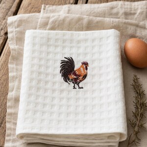 Può includere: Un asciugamano da cucina bianco piegato, a trama a nido d'ape, con un disegno di gallo ricamato in marrone, arancione e nero. L'asciugamano è su un panno di lino beige, con un uovo marrone e un rametto di rosmarino. Sfondo in legno.