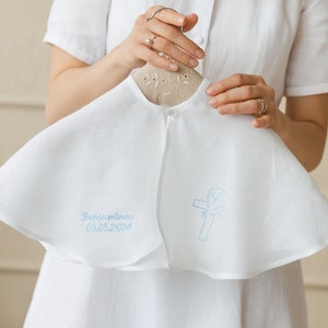 May include: White christening gown with a blue embroidered cross and dove design. The gown has a ruffled hem and a button closure at the neck. The text "Benjaminas 03.05.2020" is embroidered on the gown.
