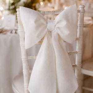 Puede incluir: Una silla blanca adornada con un gran lazo decorativo de tela suave. El lazo es el elemento central, añadiendo un toque de elegancia. La silla forma parte de un conjunto, adecuado para una boda o una ocasión formal.