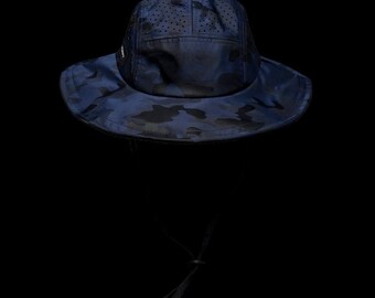 May include: A dark blue camouflage bucket hat featuring a perforated crown for ventilation and a wide brim. The hat includes an adjustable chin strap with a sliding toggle, providing a secure fit. The design displays a subtle tonal pattern.