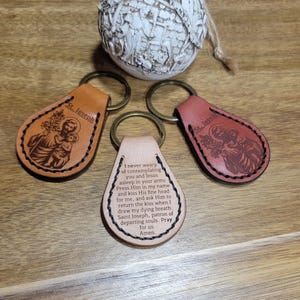 Puede incluir: Tres llaveros de cuero con anillos de latón. Dos llaveros son marrones y rojos, con una imagen de San José y un niño. El tercer llavero es de color beige con una oración inscrita. Una esfera decorativa está en el fondo.