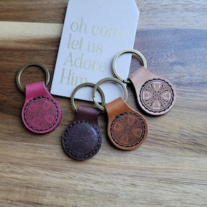 Peut inclure: Quatre porte-clés en cuir dans les tons de rose, de marron et de beige, chacun avec un motif circulaire et un anneau de couleur laiton. Les porte-clés ont un motif de nœud celtique. Une étiquette avec les mots "oh come let us Adore Him" est en arrière-plan.