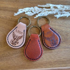 Op de afbeelding: Drie leren sleutelhangers met messing ringen. Elke sleutelhanger is druppelvormig en heeft de tekst "St. Michael the Archangel" en een afbeelding van een engel. De sleutelhangers zijn in tinten beige, roodbruin en bruin.