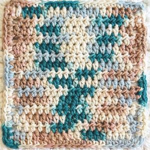 May include: A square crochet coaster or washcloth featuring a variegated yarn pattern in shades of teal, cream, light blue, and dusty rose. The textile displays a consistent, dense stitch pattern throughout, creating a textured surface.