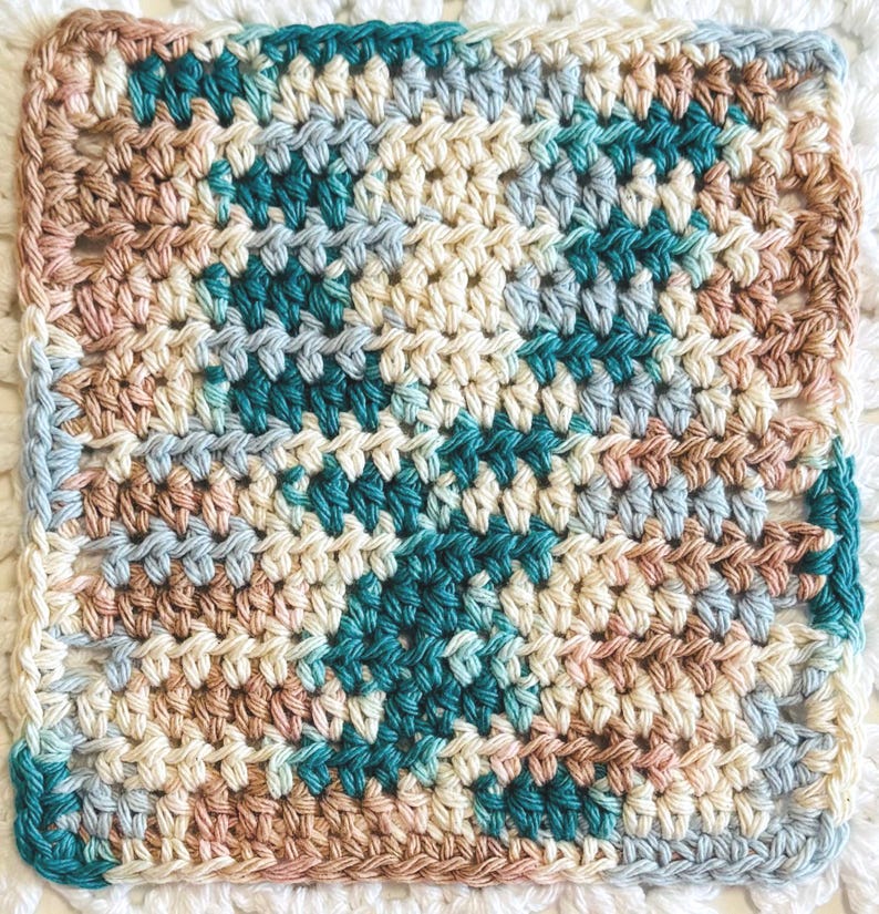 May include: A square crochet coaster or washcloth featuring a variegated yarn pattern in shades of teal, cream, light blue, and dusty rose. The textile displays a consistent, dense stitch pattern throughout, creating a textured surface.