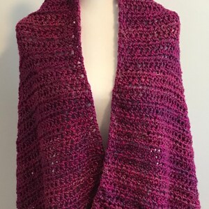 Op de afbeelding: Een gehaakte sjaal in een diepe paarse kleur met een subtiel ombre-effect. De sjaal is over een wit mannequin gehangen.
