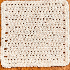 May include: A square-shaped, off-white crochet washcloth featuring a consistent, repeating textured stitch pattern throughout the entire surface. The edges are finished with a simple, uniform border that frames the square design neatly.