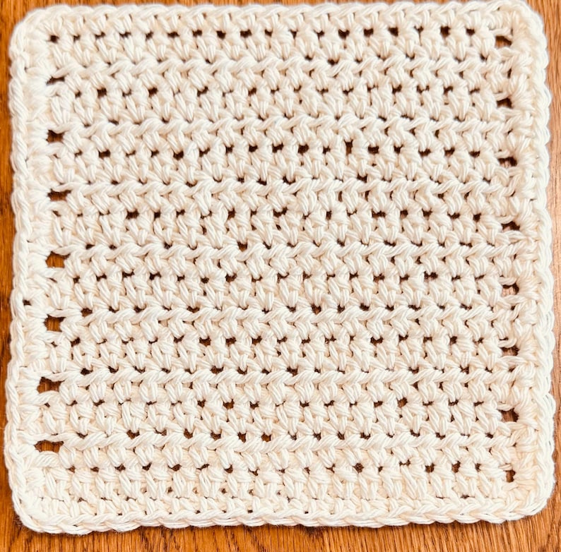 May include: A square-shaped, off-white crochet washcloth featuring a consistent, repeating textured stitch pattern throughout the entire surface. The edges are finished with a simple, uniform border that frames the square design neatly.
