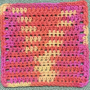 May include: A square crochet coaster featuring a variegated yarn pattern in shades of bright pink, orange, and pale yellow. The textile displays a consistent, dense stitch texture throughout, creating a uniform surface with a repeating color gradient.