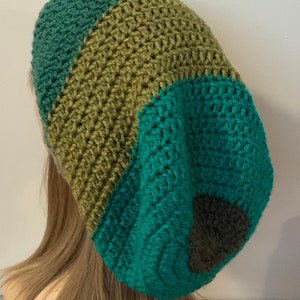 May include: A handmade crochet hat in shades of teal, olive green, and gray. The hat features a striped design with a rounded shape, perfect for cold weather. The hat is made of soft yarn and is a stylish accessory.