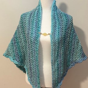 May include: A crocheted shawl in shades of blue and green, with a scalloped edge. The shawl is draped over a white mannequin.