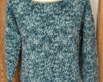 Op de afbeelding: Een teal en blauw gespikkelde trui met een ronde hals en lange mouwen. De halslijn en zoom zijn afgewerkt met een dunne, lichtgouden rand. De trui wordt getoond op een witte mannequin, die de textuur van de gebreide stof benadrukt.