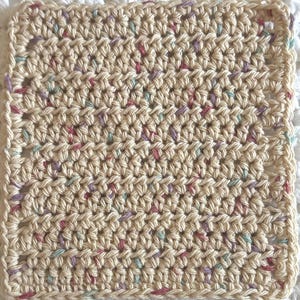 May include: A square crochet coaster made of cream-colored yarn with small, scattered flecks of pink, purple, and light blue throughout. The piece features a consistent, textured stitch pattern with a simple border along all four edges.