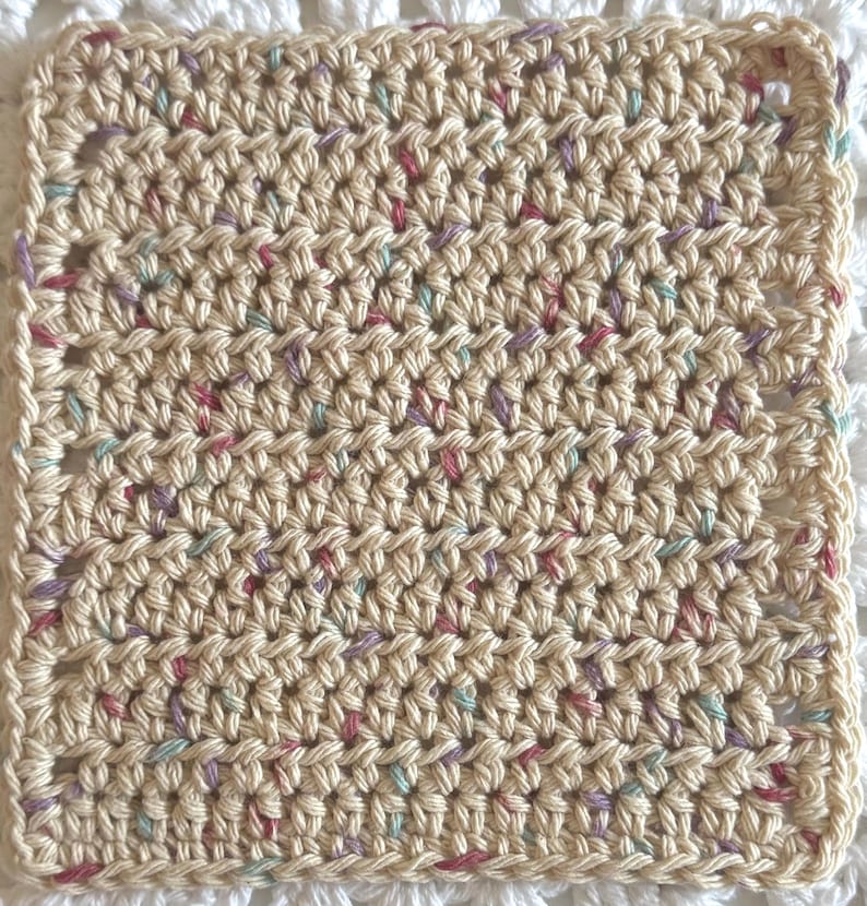 May include: A square crochet coaster made of cream-colored yarn with small, scattered flecks of pink, purple, and light blue throughout. The piece features a consistent, textured stitch pattern with a simple border along all four edges.