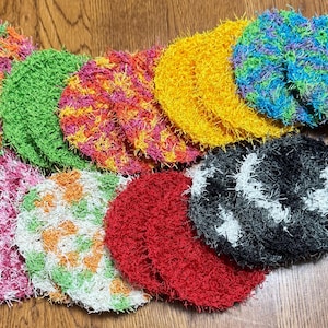 Puede incluir: Una colección de estropajos redondos y coloridos, tejidos a ganchillo en varios colores, incluyendo rosa, verde, amarillo, azul, rojo y negro. Los estropajos están dispuestos sobre una superficie de madera, mostrando su aspecto texturizado y hecho a mano.