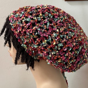 May include: A crochet hat featuring a multi-colored textured yarn in shades of pink, teal, yellow, and black. The hat has an open, loose-knit construction and is worn over dark hair, covering the crown of the head with a rounded, slouchy shape.