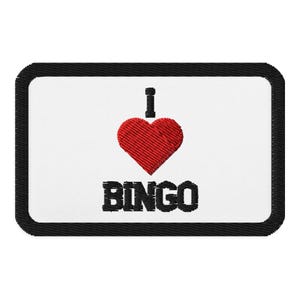 Puede incluir: Parche blanco bordado con borde negro. El parche presenta un corazón rojo con el texto "Me encanta el BINGO" en negro.