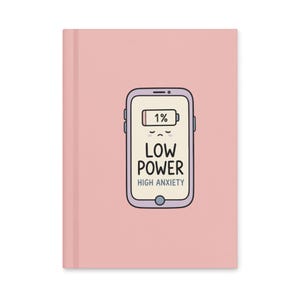 May include: A light pink hardcover notebook featuring a central illustration of a smartphone with a purple border. The screen displays a 1% battery icon, a sad face, and the text LOW POWER HIGH ANXIETY in black and blue lettering.