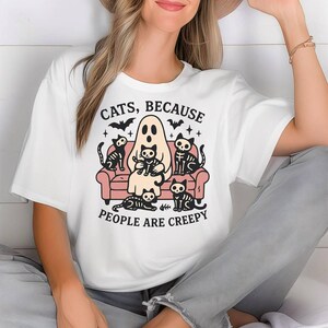 May include: White t-shirt with a Halloween graphic. The design shows a ghost on a pink sofa with skeleton cats. The text reads "CATS, BECAUSE PEOPLE ARE CREEPY" in black, with bats and stars. A fun, spooky design.